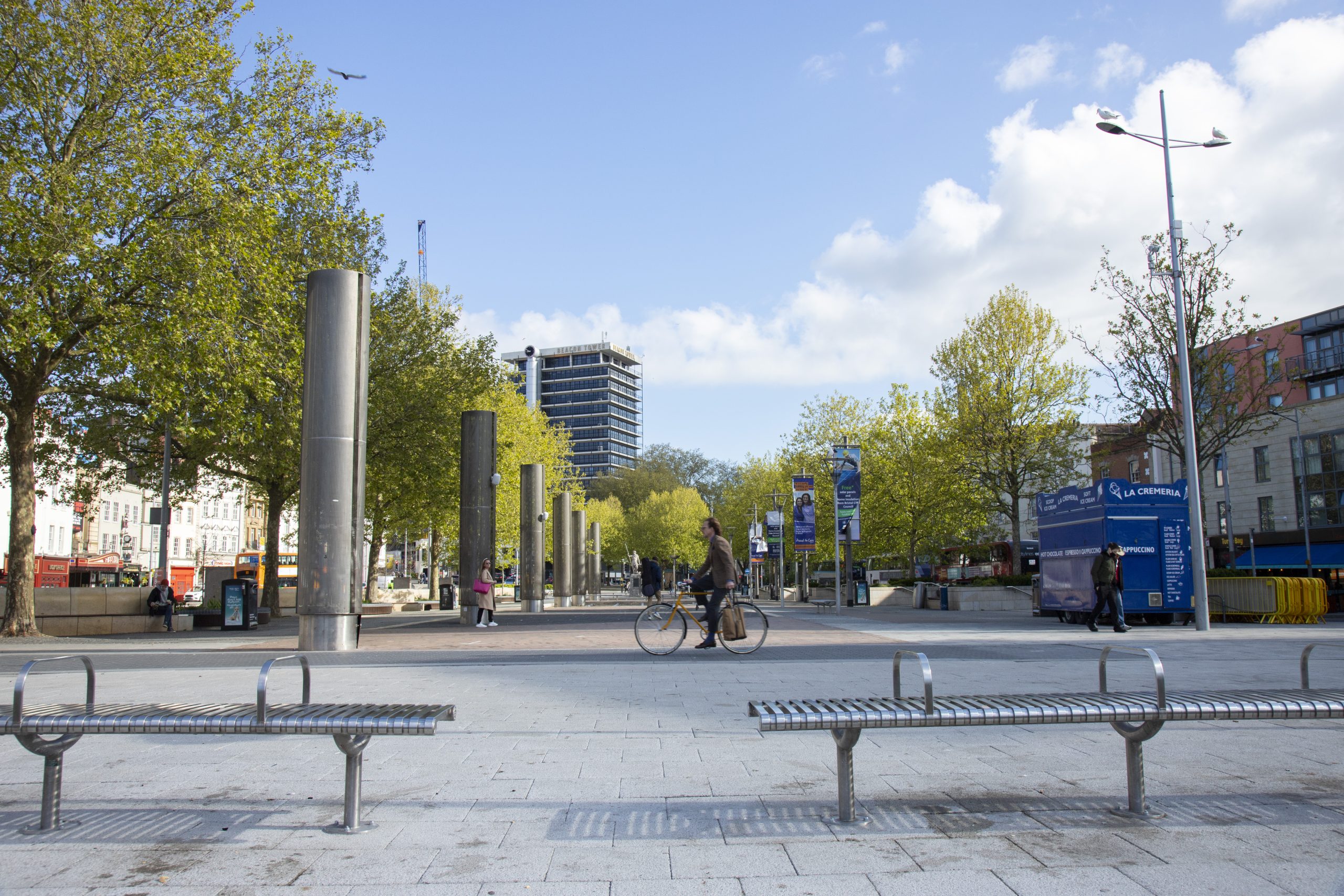 Bristol City Centre view with trees, buildings, benches and members of the public.